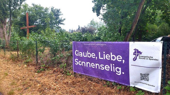 Lila Banner am Zaun: Glaube, Liebe, Sonnenselig.