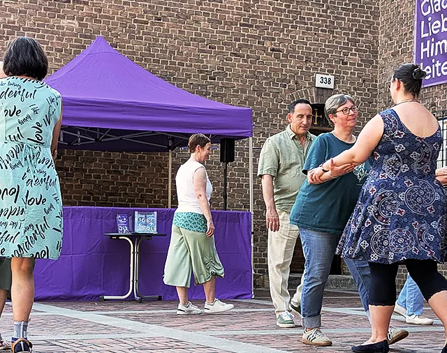 Paare tanzen Tango auf dem Kirchvorplatz, Banner Glaube, Liebe, Himmelsstürmer hängt an Backsteinwand