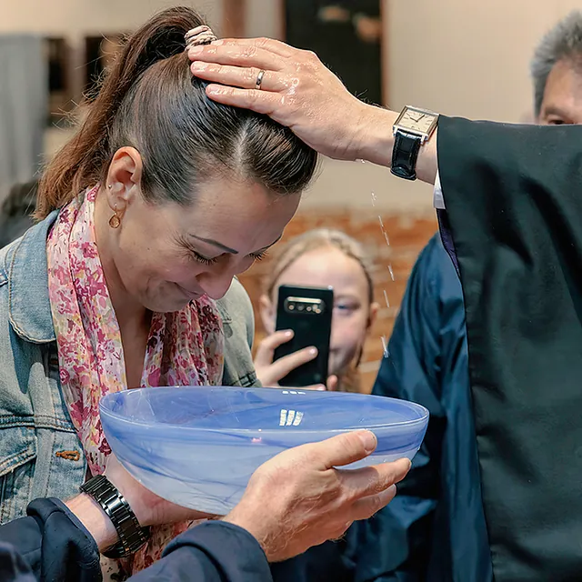Pfarrer tauft Frau, ein Mann hält die Taufschale hoch