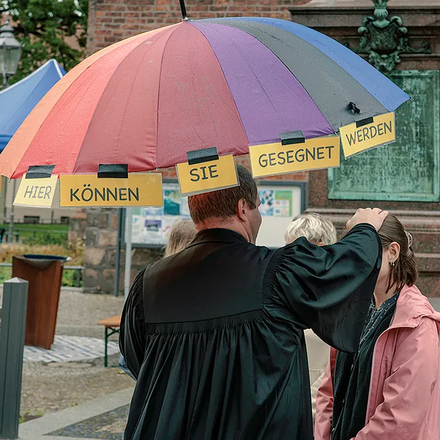 Pfarrer mit Regenbogenschirm segnet eine Frau