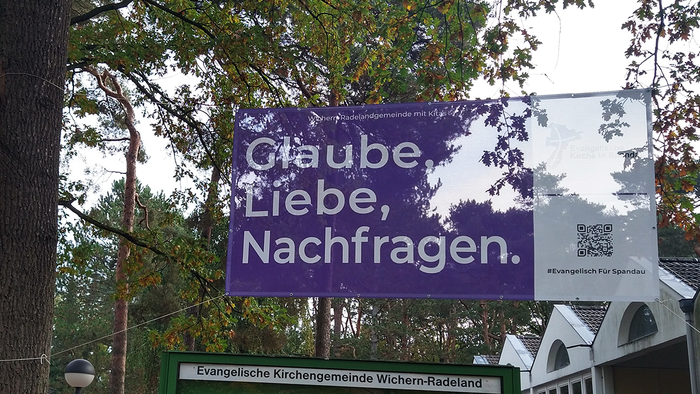 Lila Banner zwischen Bäumen: Glaube, Liebe, Nachfragen.