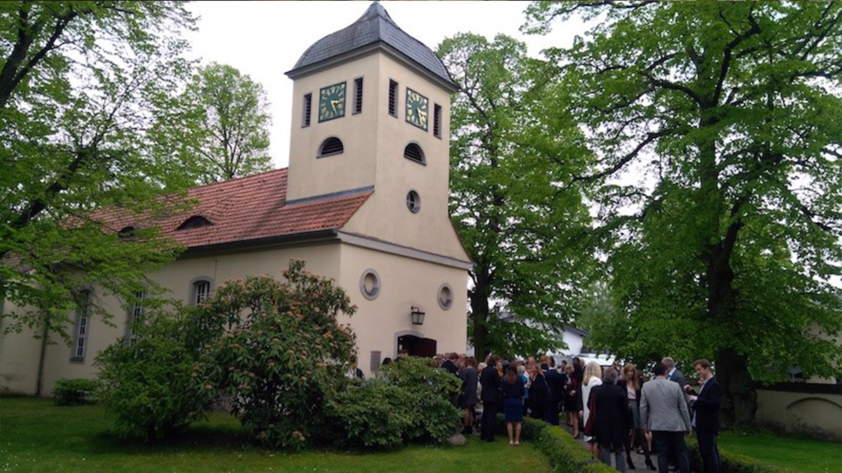 Dorfkirche Kladow umgeben von grünen Bäumen. Vor der Kirche stehen festlich gekleidete Menschen.