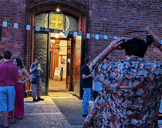 DJ von hinten, Personengrüppchen und erleuchteter Eingang der St. Nikolaikirche am Abend