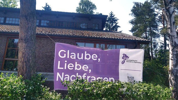 Lila Banner zwischen Bäumden: Glaube, Liebe, Nachfragen.