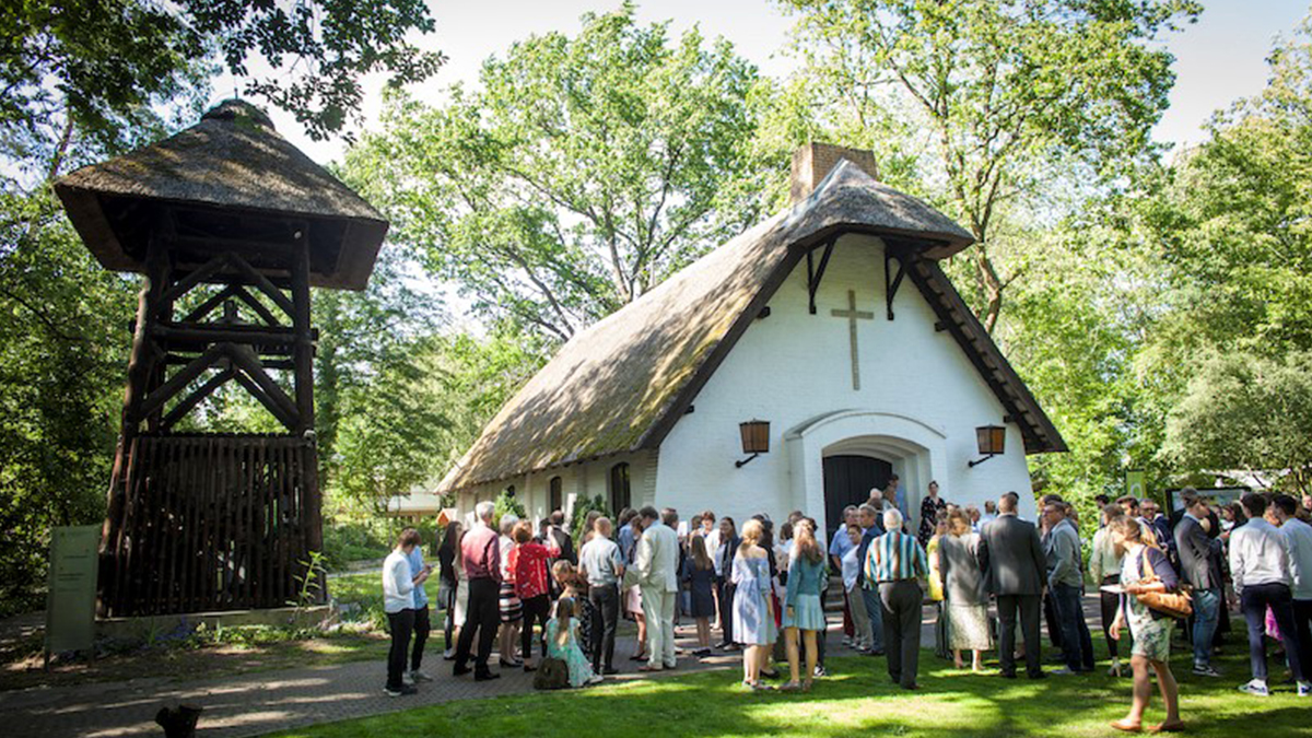 Schilfdachkapelle umgeben von grünen Bäumen. Vor dem Gebäude stehen Menschen in Grüppchen.