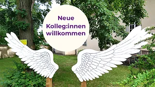 Schild "Neue Kolleg:innen willkommen" zwischen zwei Engelsflügeln.