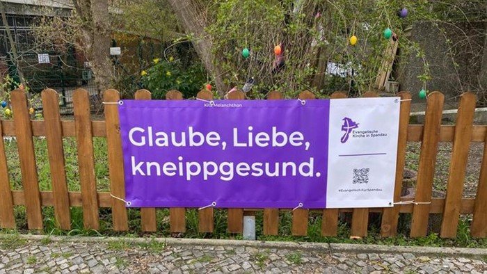 Lila Banner am Zaun: Glaube, Liebe, kneippgesund.