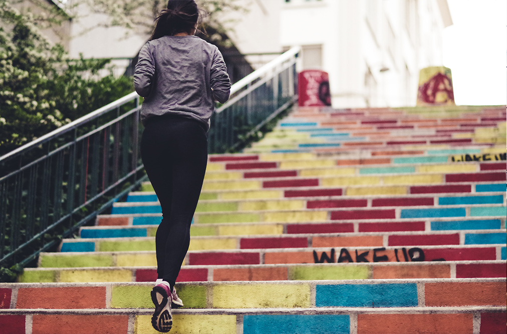 Ein Frau joggt eine Treppe hinauf, deren Stufen bunt angemalt sind.
