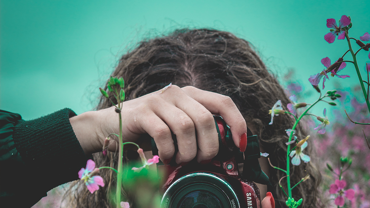 Eine Frau fotografiert mit einer Kamera zwischen Blumen hindurch.