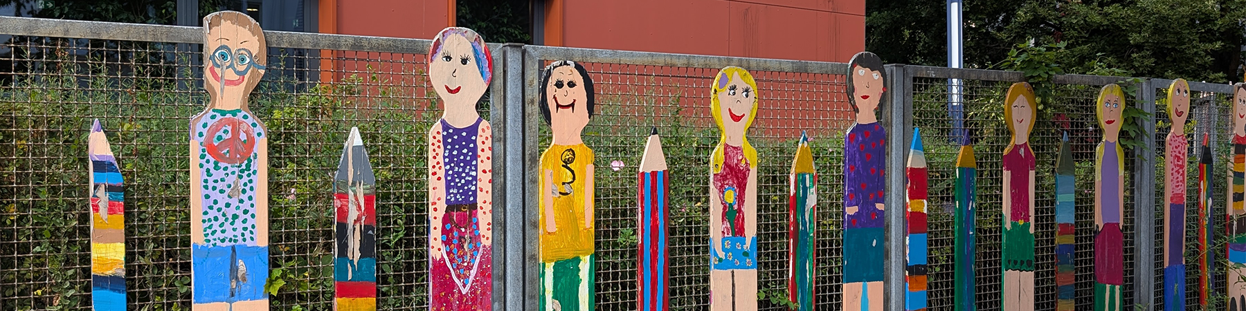 Zaun vor der Grundschule mit bunt bemalten Kinderfiguren und Stiften