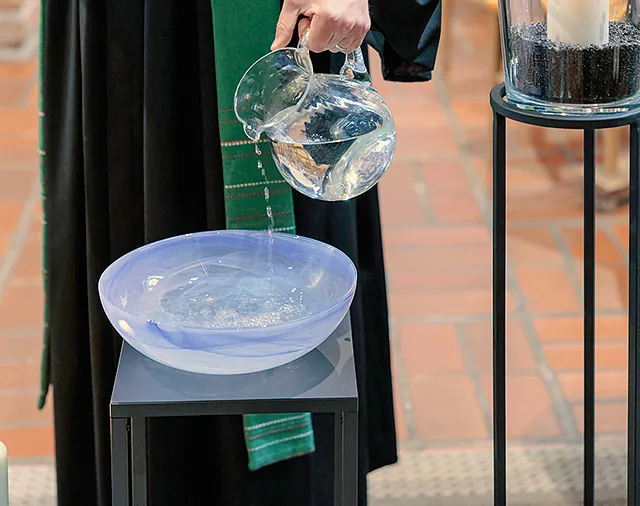Pfarrer gießt mit Glaskrug Wasser in blaue Taufschale