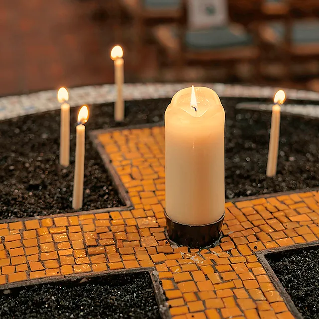 Kerzentisch aus gelben Mosaik und großer Kerze in der Mitte, im schwarzen Sand stecken kleine Kerzen