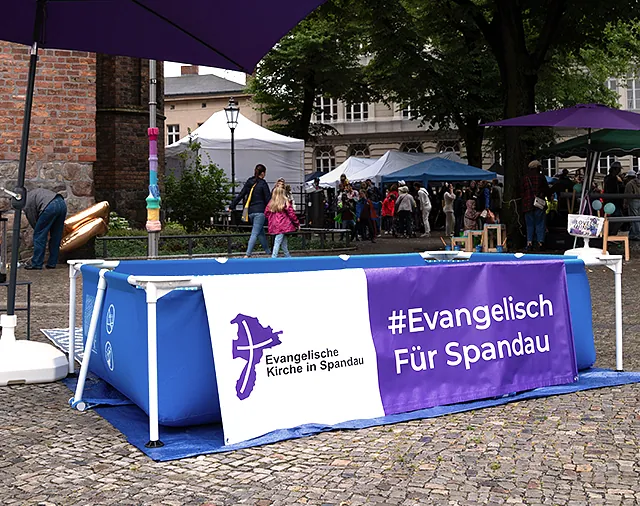 Wasserbecken mit Banner #Evangelisch für Spandau, dahinter Menschen vor und unter Pavillons