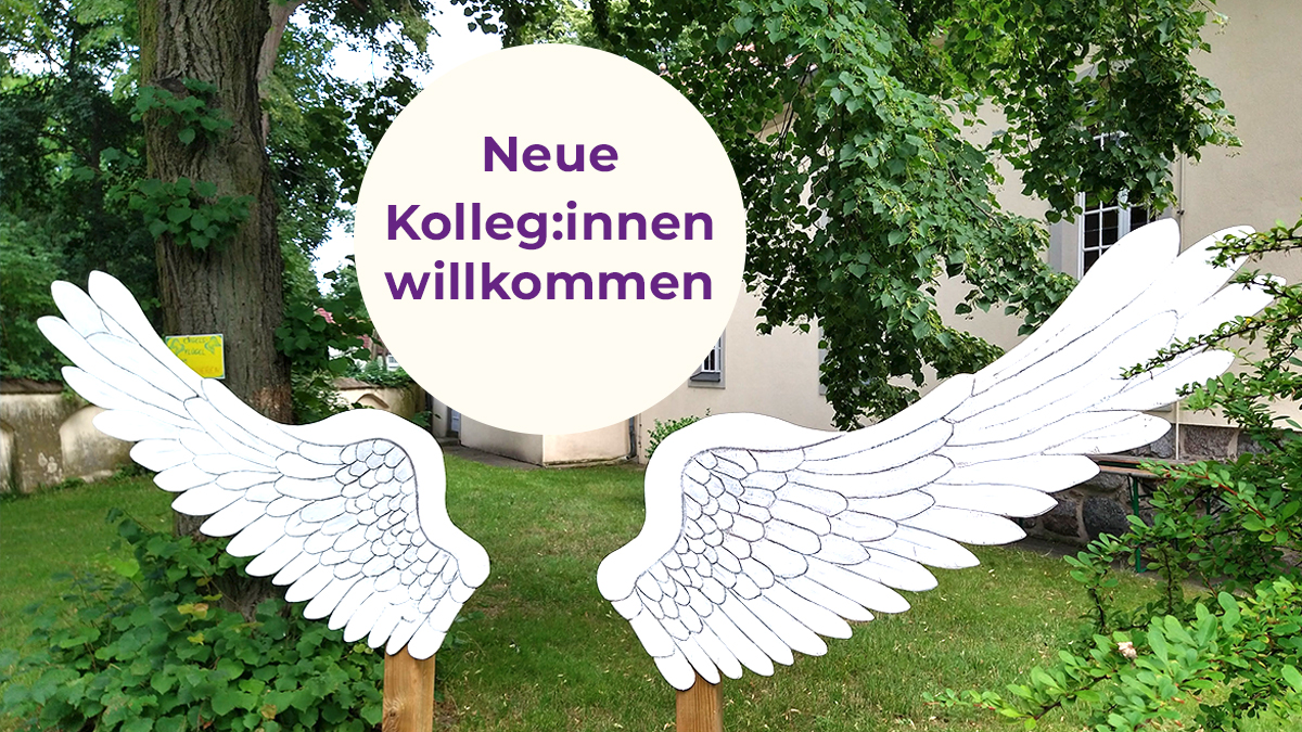 Ein Schild "Neue Kolleg:innen willkommen" zwischen zwei hölzerne Engelsflügel im Pfarrgarten.